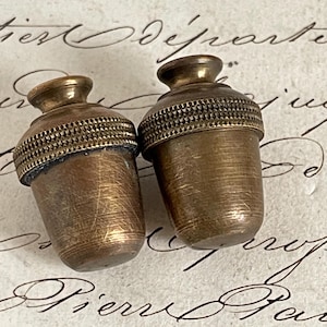 May include: Two antique brass acorn-shaped objects. Each has a textured band around the middle and a small, rounded top. The objects are set against a backdrop of aged paper with handwritten script.