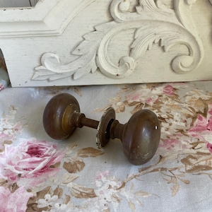 May include: A vintage brass door knob set featuring two rounded, weathered brass knobs connected by a central metal spindle and a single circular metal rosette plate. The hardware displays a tarnished, aged patina across its metallic surface.