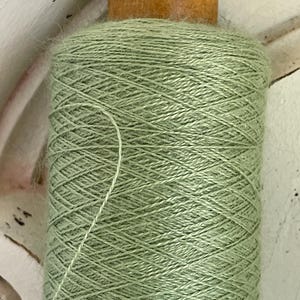 May include: A spool of sage green yarn with a wooden top. The yarn is tightly wound, creating a textured surface. A strand of yarn extends from the spool. The background is a distressed white surface.