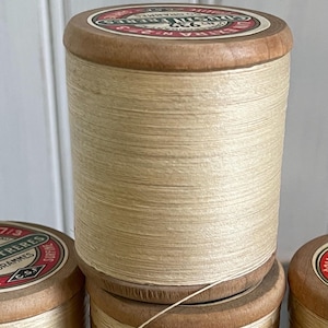 Puede incluir: Primer plano de varios carretes de hilo beige. Los carretes tienen tapas y fondos de madera con etiquetas rojas y blancas. El hilo está enrollado firmemente alrededor del carrete. Los carretes están apilados. El hilo es de color claro y neutro.