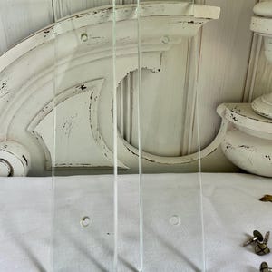 Puede incluir: Tres expositores de vidrio transparente con forma rectangular y un pequeño agujero. Los expositores están colocados frente a un elemento arquitectónico decorativo blanco y desgastado. El fondo incluye una superficie blanca y otros objetos decorativos.
