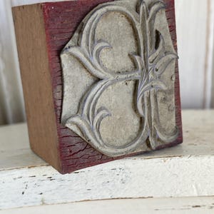 Może przedstawiać: Zabytkowy drewniany klocek do druku z literą "E" w ozdobnej czcionce. Litera jest w kolorze jasnoszarym, z bogatym wzorem. Klocek ma czerwoną powierzchnię i podstawę z naturalnego drewna.