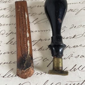 May include: A black wax seal stamp with a rounded handle and a brass collar, positioned next to a rectangular stick of copper-colored sealing wax that features embossed text and a worn, textured end with signs of previous use.