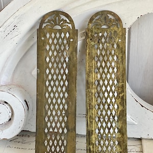 Puede incluir: Dos cubiertas de ventilación antiguas de latón. Cada cubierta rectangular presenta un patrón de celosía de diamantes y una parte superior arqueada con un diseño floral decorativo. El latón tiene una pátina cálida y envejecida.