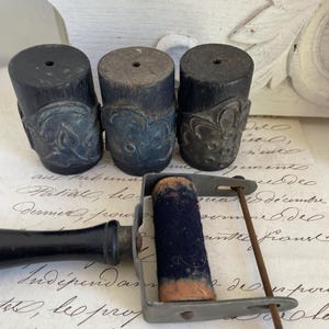 May include: Three cylindrical dark wood printing blocks with carved relief patterns and a metal ink roller tool featuring a black handle and a fabric-covered cylinder. The printing blocks have flat tops with small central holes and intricate designs.
