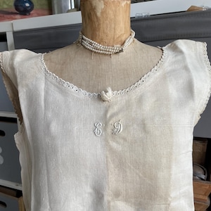 May include: A sleeveless off-white linen garment featuring a scalloped lace trim along the neckline and armholes. The centre of the chest displays embroidered initials in a cursive script, and a small fabric bird ornament is attached at the neckline.