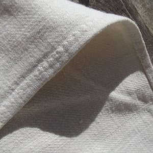 May include: Close-up of a folded, off-white linen fabric. The fabric has a textured weave and a visible seam along the edge. The natural fibers create a soft, neutral aesthetic. The fabric is likely used for home decor or apparel.