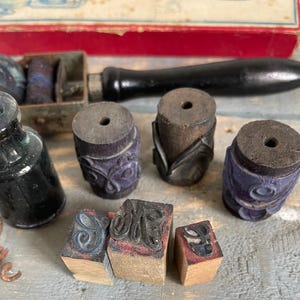 Pode incluir: Uma variedade de ferramentas de impressão vintage, incluindo carimbos de madeira com letras e desenhos decorativos, almofadas de tinta e um frasco de tinta preta. Os carimbos têm uma base de madeira e uma superfície de carimbo de borracha. O frasco de tinta é feito de vidro escuro.