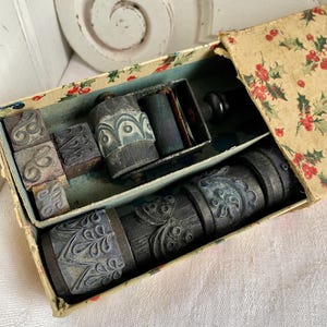 May include: A vintage printing set in a box with a floral pattern. The set includes several decorative rollers and individual letter stamps. The rollers and stamps are dark gray with ornate designs. The box is open, revealing the contents.