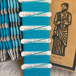 May include: A stack of turquoise Laine St. Pierre thread spools, each labeled with text in French. The spools are stacked vertically, with a vintage illustration of a saint holding a key in the background. The text on the spools reads "4 fils de 4 mètres = 16 mètres".