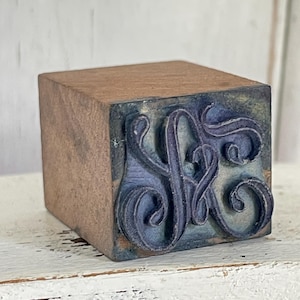 Puede incluir: Un sello de bloque de madera con un diseño de monograma azul oscuro. El sello presenta una letra "A" adornada y estilizada con remolinos decorativos. El bloque de madera tiene un acabado marrón natural, sobre un fondo blanco.