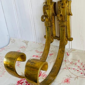 May include: Pair of antique brass wall sconces with ornate detailing. The sconces have a curved design with a circular opening at the bottom. The brass has a warm, aged patina. The background is a floral patterned fabric.