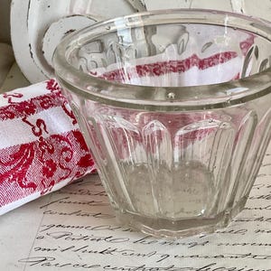 May include: A clear, vintage glass bowl with a fluted design. The bowl sits on a surface with handwritten script and is next to a red and white patterned cloth. The bowl has a thick rim and a slightly flared shape.