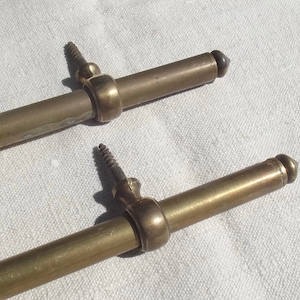 May include: Two brass curtain rods with decorative finials. The rods are approximately 45 cm long and have a diameter of about 1.25 cm.