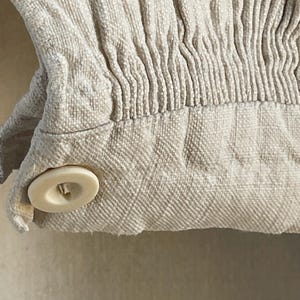 以下が含まれることがあります： オフホワイトの丸いボタンが1つ付いたベージュの生地の袖口で、縦方向のスモッキング加工が施されています。素材には目に見える織り目があり、袖口はボタンで留められており、裾の上にあるギャザーのひだが見えるデザインになっています。