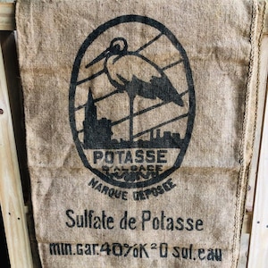 May include: A burlap sack with a black and white logo of a stork standing on a building. The text "Potasse Marque Deposee" is printed in a circle around the stork. The text "Sulfate de Potasse min.gar.40%K2O sul.eau" is printed below the logo.