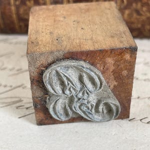 May include: A vintage wooden stamp with a decorative metal design. The stamp has a rectangular wooden block with a detailed, possibly floral, metal imprint on one side. The wood shows signs of age and wear.