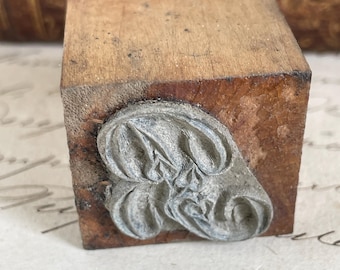 May include: A vintage wooden stamp with a decorative metal design. The stamp has a rectangular wooden block with a detailed, possibly floral, metal imprint on one side. The wood shows signs of age and wear.