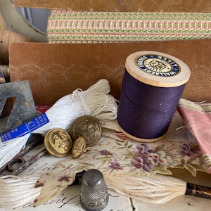Puede incluir: Una colección de suministros de costura vintage, que incluye una bobina de hilo morado oscuro, hilo blanco, botones dorados, un dedal y tela floral. También se ven cajas de cuero marrón y adornos decorativos.
