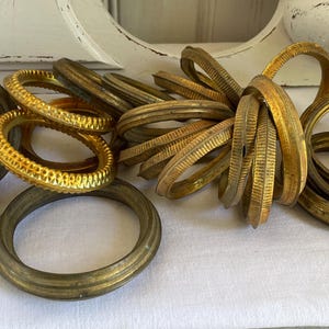 Puede incluir: Una colección de anillos de latón antiguos de varios tamaños y texturas. Algunos anillos son lisos, mientras que otros tienen patrones detallados. Los anillos son en tonos dorados y bronce, lo que sugiere edad y uso. Los artículos están dispuestos sobre una superficie blanca.