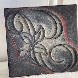May include: A square, wooden block with a carved floral design. The design features swirling lines and leaf shapes in a light gray color, set against a dark gray background with a red border. The block is likely used for printing or stamping.