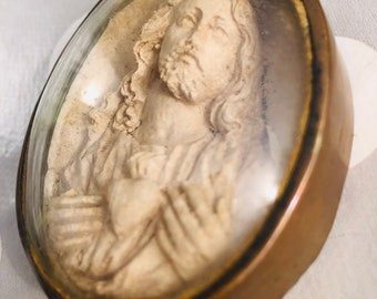 May include: Oval-shaped religious pendant featuring a bas-relief of a figure with long hair and a beard, framed by a clear cover and a bronze-colored metal casing. The figure is depicted with hands clasped near the chest. The pendant has a vintage aesthetic.