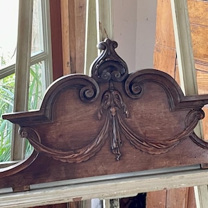 May include: Ornate, dark brown wooden architectural element with carved details. Features a central floral design, scrolling edges, and finials. The piece is likely a decorative accent for furniture or a building.