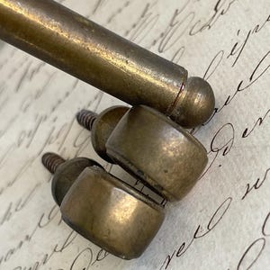 Puede incluir: Primer plano de herrajes de muebles de latón antiguo, que incluyen un asa cilíndrica y varios pomos pequeños roscados. El herraje tiene un aspecto deslustrado y envejecido, lo que sugiere piezas antiguas o recuperadas. El fondo es una hoja de papel con escritura manuscrita.