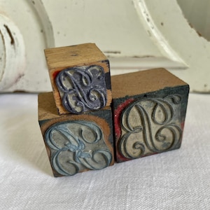 Könnte beinhalten: Drei Vintage-Holzbuchstabenstempel mit verzierten Buchstabenmustern. Die Stempel zeigen den Buchstaben "B" in einer dekorativen, wirbelnden Schriftart. Die Stempelkissen weisen Spuren von blauer, grauer und schwarzer Tinte mit roten Rändern auf.