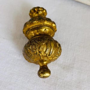 May include: A gold-colored, ornate decorative object with a bulbous shape. The object has a textured surface and a small, round base. The top features a floral-like design. The overall design is reminiscent of antique hardware.
