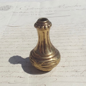 Op de afbeelding: Een vintage messing flesstopper met een sierlijk ontwerp. De stopper heeft een bolvormige basis met horizontale groeven en een gegroefde hals, bekroond met een decoratieve rand. Het staat tegen een achtergrond van verouderd papier met vervaagde tekst.