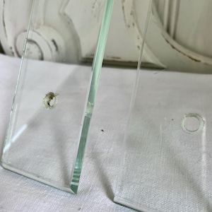 May include: Two clear glass panels with a beveled edge and a small hole. The panels are slightly transparent, allowing a glimpse of the background. The glass has a smooth, reflective surface.