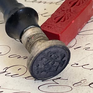 Könnte beinhalten: Ein Wachssiegelstempel mit schwarzem Griff und einem silberfarbenen Metallkopf mit eingraviertem Wappenmotiv. Neben dem Stempel liegt eine rechteckige rote Wachsstange mit einem dekorativen Prägemuster auf der Oberfläche, die auf Papier liegt.