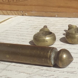 May include: Antique brass objects, including two small ring-shaped pieces and a long, cylindrical item with a rounded end. The items are arranged on a sheet of aged paper with handwritten script, suggesting a vintage aesthetic.