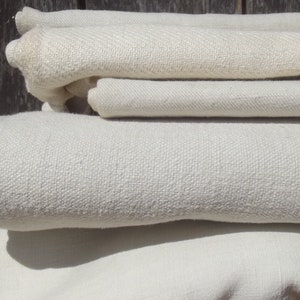 Puede incluir: Una pila de cuatro telas de lino de color blanco roto. Las telas están dobladas y apiladas una encima de la otra. La tela tiene una textura sutil.