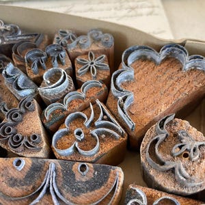 Puede incluir: Una colección de sellos de madera vintage con varios diseños, incluyendo flores, corazones y formas abstractas. Los sellos tienen contornos metálicos y están colocados en una caja de cartón. La madera tiene un tono marrón cálido.