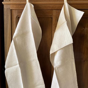 Puede incluir: Dos toallas de lino color crema colgadas de ganchos de madera. Las toallas están dobladas y drapeadas, mostrando su superficie texturizada y color neutro. El fondo es un armario de madera oscura.