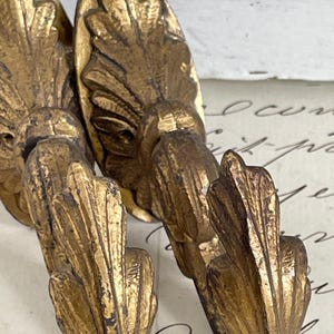 Op de afbeelding: Close-up van verschillende antieke goudkleurige wandhaken. De haken hebben een gedetailleerd, sierlijk ontwerp met een bladachtige vorm. De haken zijn gemaakt van metaal en hebben een verweerde afwerking, waardoor ze een vintage uitstraling hebben.