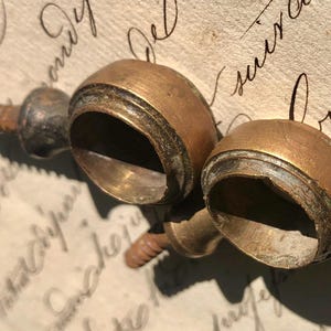 May include: Two antique brass furniture knobs with threaded posts. The knobs have a rounded, cup-like shape with a dark, aged patina. The background features faded script writing.