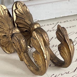 Puede incluir: Ganchos de pared de estilo antiguo con acabado dorado. Los ganchos presentan un diseño detallado en forma de hoja. Probablemente son de metal y adecuados para colgar objetos. Montados sobre una superficie blanca.