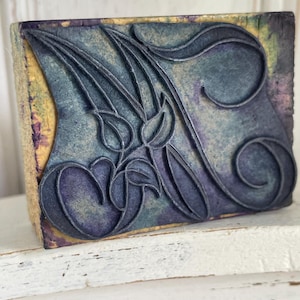 Puede incluir: Un sello rectangular de estilo vintage con un diseño floral. El sello presenta un patrón floral estilizado en relieve en tonos azules y morados sobre un fondo texturizado. El bloque de madera tiene un aspecto desgastado y envejecido, con toques de amarillo y morado.