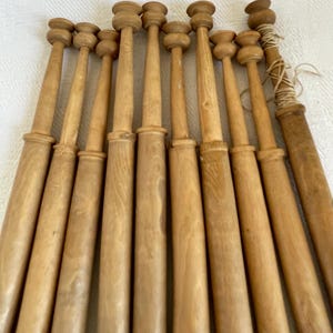 Può includere: Una collezione di dieci bobine in legno marrone chiaro disposte in fila. Ogni bobina presenta un lungo fusto cilindrico con una parte superiore arrotondata e svasata e una sezione a colletto distinta. Una bobina all'estrema destra ha del filo chiaro avvolto.