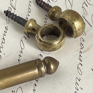 May include: Close-up of three antique brass hardware pieces. Two pieces have a ring shape with a screw, and the third is a cylindrical shape with a rounded tip. The hardware is aged with a vintage aesthetic.