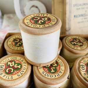 Pode incluir: Uma caixa de carretéis de linha vintage J & P Coats. Os carretéis são de madeira e têm um rótulo branco com um trevo verde e o texto "J & P. Coats 500 Yds 406 Fils".