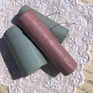 May include: Two rolls of paper, one light blue and one pink with white polka dots, are tied together with twine. The rolls are on a white lace doily.