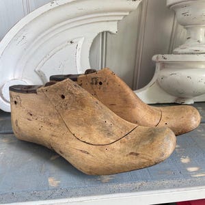 May include: Two wooden shoe lasts, one on top of the other, with a light brown color and a worn, aged appearance. The lasts have a rounded toe and a slight heel.