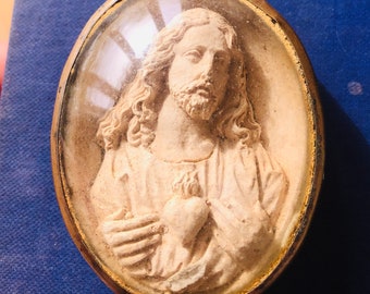 Op de afbeelding: Ovale religieuze hanger met een bas-reliëf van Jezus Christus. De afbeelding is ingekapseld in helder, gewelfd glas, gezet in een aangetaste goudkleurige metalen lijst. De achtergrond is diepblauw.