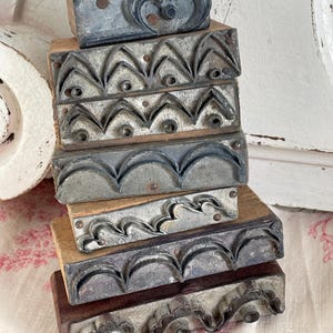 May include: A stack of antique printing blocks with various decorative metal designs mounted on wooden bases. The blocks feature floral, geometric, and curved patterns in shades of gray and silver, showing signs of age and use.
