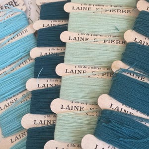 May include: A collection of vintage Laine St. Pierre yarn spools in various shades of blue and green. Each spool is labeled with text indicating the yarn type and length. The spools are arranged in neat rows, showcasing the different colors.