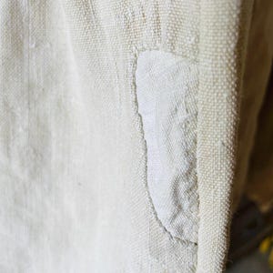 May include: Close-up of a white linen fabric with a sewn-in patch. The fabric has a textured weave and the patch is slightly raised.